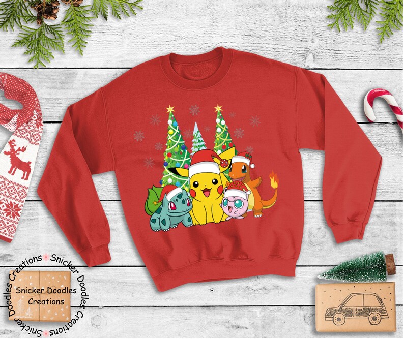 May include: Red Christmas sweater with a graphic of Pikachu, Bulbasaur, Charmander, and Jigglypuff standing in front of two Christmas trees. The Pokemon are wearing Santa hats. The sweater has the words 'Snicker Doodles Creations' printed on the bottom left and bottom right.