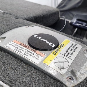 May include: A close-up of a gray metal boat seat base with a black LUND logo sticker and two warning labels. The label on the left reads "AVOID SERIOUS OR FATAL INJURY. DO NOT OCCUPY SEAT WHEN SPEED EXCEEDS 5 MPH." The label on the right reads "CAUTION DO NOT TRIM ENGINE WITH A SEAT INSTALLED AT THIS LOCATION."