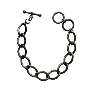 May include: A dark gray metal bracelet with oval links and a toggle clasp. The bracelet is made of a series of interconnected oval-shaped links, with a circular link at the end. The toggle clasp is a T-bar design.