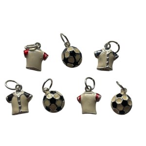 May include: A collection of silver-toned soccer-themed charms. The charms include miniature soccer balls and soccer jerseys in cream, black, and red. The charms are designed for jewelry making or crafting.