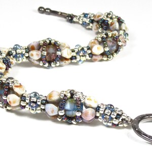 以下が含まれることがあります： 銀色のリング留め具が付いたビーズブレスレット。ブレスレットは、青、白、茶色の虹色をした小さなビーズで作られています。ビーズはらせん状に配置されています。