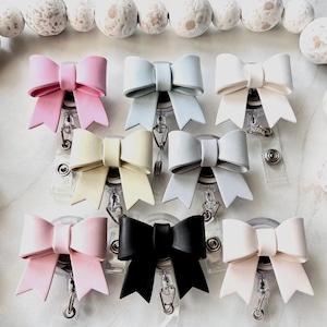 May include: A collection of bow-shaped badge reels in various colors, including pink, light blue, white, yellow, gray, and black. Each bow is attached to a clear plastic reel with a metal clip. Decorative elements are visible in the background.