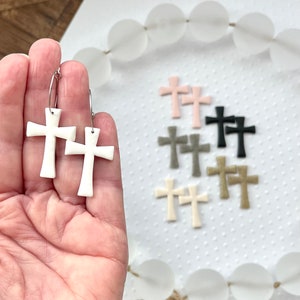 May include: White ceramic cross hoop earrings with silver hoops. The earrings are shown in a hand. There are also multiple pairs of cross earrings in other colors in the background, including pink, black, gray, and gold.