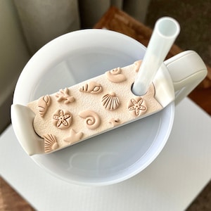May include: A white tumbler with a beige lid featuring a variety of sea-themed shapes, including seashells, starfish, and a nautilus shell. A clear plastic straw is inserted through the lid.