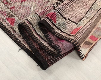 May include: A rolled-up, textured rug with a geometric pattern. The rug features a color palette of pink, gray, and black, with a border of dark brown and black. The design includes shapes like triangles and squares.