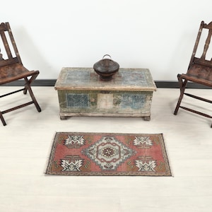 May include: An antique wooden chest with a metal handle sits between two folding chairs. A small, patterned rug in shades of red, brown, and white is on the floor. The chairs and chest are made of dark wood.