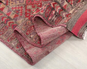 May include: A folded, rectangular rug with a geometric pattern in shades of red, pink, and gray. The rug's edges are a darker shade of pink, and the design includes a striped border. The rug is made of a woven material.