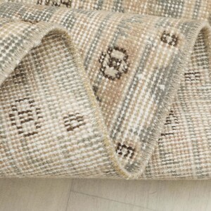 May include: Close-up of a rolled-up, vintage-style rug. The rug features a beige, gray, and brown color palette with a geometric pattern. The edges are frayed, giving it a distressed look. The rug's texture is visible, suggesting a woven construction.