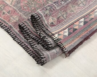 May include: Close-up of a folded rug with a black fringe. The rug has a pink, gray, and white geometric pattern.