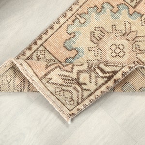 Puede incluir: Una alfombra rectangular plegada con un aspecto envejecido y vintage. La alfombra presenta un patrón geométrico repetitivo en tonos beige, rosa claro y azul, con acentos marrones. Los bordes están deshilachados.