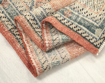 May include: A folded, rectangular rug with a geometric pattern. The rug features a color palette of coral, teal, cream, and black. The edges are finished with a coral border. The rug appears to be made of woven material.