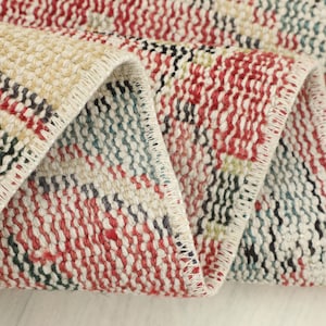 May include: Close-up of a folded, rectangular rug with a woven texture. The rug features a patchwork design with red, cream, black, green, and blue hues. The edges are neatly finished with a cream-colored border.