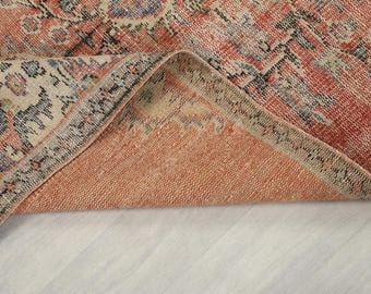 May include: A folded rug with a distressed appearance and a complex pattern. The rug's colours include coral, beige, and blue, with a visible border. The folded section reveals the underside, which is a solid coral colour. The rug is likely made of woven material.
