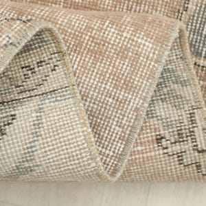 May include: Close-up of a rolled-up, vintage-style rug. The rug features a distressed, geometric pattern in shades of beige, brown, and gray. The edges are frayed, adding to the rug's antique aesthetic. The rug is likely made of woven material.