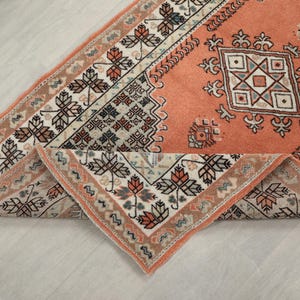 May include: A peach-coloured rug with a geometric pattern in shades of white, grey, and brown. The rug has a fringe border and is folded in half.