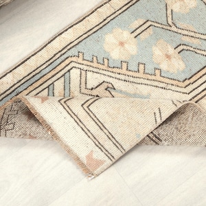 May include: A folded, vintage-style rug with a light blue central field featuring floral patterns. The rug has a beige border with geometric designs and dark brown accents. The rug's edges are frayed, giving it a worn appearance.