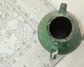 Può includere: Un vaso in ceramica verde usurato con due manici e un'ampia apertura. Il vaso è posto su un tappeto bianco sporco a fantasia. Il vaso presenta un aspetto invecchiato e rovinato.
