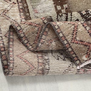 Pode incluir: Close-up de um tapete marrom e rosa com uma borda desfiada. O tapete está dobrado ao meio, revelando o design intrincado tecido.