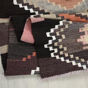 May include: A folded, woven rug with a geometric pattern. The rug features a color palette of black, brown, white, gray, and peach. The design includes zig-zag and diamond shapes. The rug's edges are frayed.
