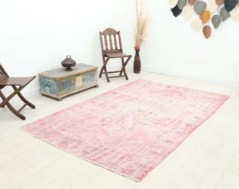 May include: A large, rectangular, distressed pink rug with a faded design. The rug is placed on a light-colored wooden floor. Two wooden chairs, a trunk, and a green vase are also in the room.
