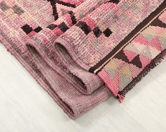 May include: A rolled-up, hand-woven rug with a pink and brown geometric pattern. The rug features a repeating design of squares and diamonds in shades of pink, brown, and beige. The edges are finished with a dark brown border and fringe.