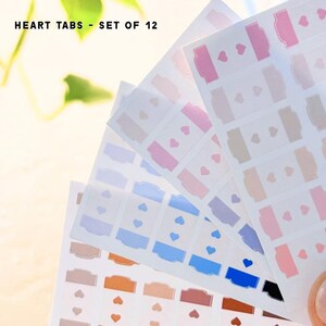 May include: A set of 12 adhesive heart tabs arranged on translucent sheets. The tabs feature various colors including pink, blue, brown, and black, each with a small heart cutout. The text "HEART TABS - SET OF 12" is printed at the top left corner.