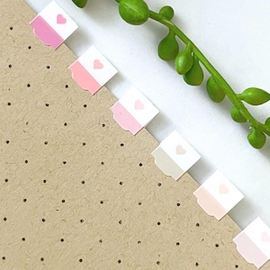 May include: A set of six square adhesive page markers attached to the edge of a tan dotted paper. Each marker features a white top section with a small pink heart and a solid colored bottom section in varying shades of pink, beige, and cream.