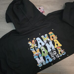 May include: Black hoodie with a hood and a colorful graphic on the back. The graphic spells "MAMA" in a playful, cartoon-like font, with each letter designed with a different pattern and color scheme. The hoodie is made of a soft material.