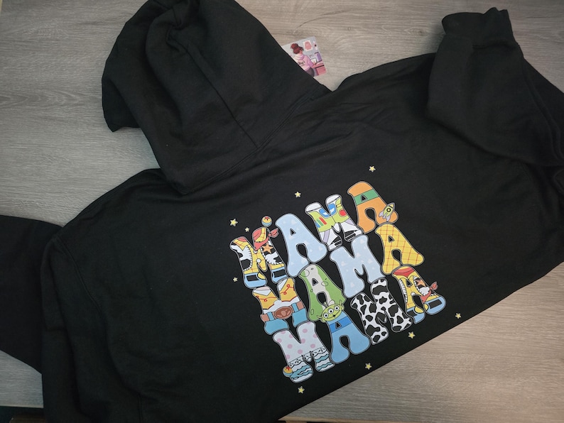 May include: Black hoodie with a hood and a colorful graphic on the back. The graphic spells "MAMA" in a playful, cartoon-like font, with each letter designed with a different pattern and color scheme. The hoodie is made of a soft material.
