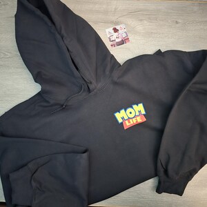 May include: Black hooded sweatshirt with the words "MOM LIFE" in a colorful, playful design on the front. The sweatshirt has long sleeves and a drawstring hood. The garment is made of a soft material.