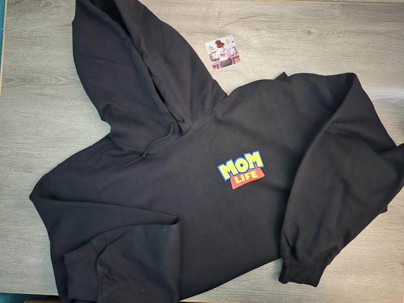 May include: Black hooded sweatshirt with the words "MOM LIFE" in a colorful, playful design on the front. The sweatshirt has long sleeves and a drawstring hood. The garment is made of a soft material.