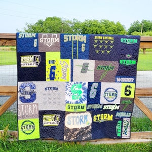 May include: A patchwork quilt made from t-shirts featuring the word "STORM" and the number "6" repeated throughout. The quilt is made in a variety of colours, including blue, green, yellow, and white.