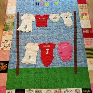以下が含まれることがあります： 青と緑のキルトに、さまざまなベビー服が縫い付けられています。キルトの中央には「Blaikyn Honey」の文字があります。ベビー服は、洗濯物干しロープに掛かっているかのように、一列に並んでいます。服には、「Biloxi」、「Sorrels 7」、「Sister」など、さまざまな言葉が書かれています。