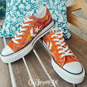 May include: Orange Converse All Star sneakers with white laces and soles. The canvas shoes feature a leaf design in red, yellow, and orange. The Converse logo is visible on the tongue and side of the shoe.