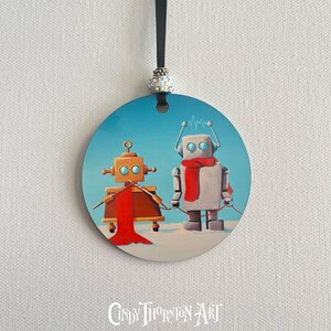 Op de afbeelding: Een ronde kerstversiering met twee robots op een besneeuwd landschap onder een blauwe lucht. Eén robot is oranje, de andere grijs, beide met ronde ogen. De oranje robot heeft een rode sjaal en de grijze robot heeft een rode sjaal. De versiering heeft een zwart lint.