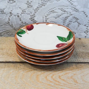 May include: A stack of five white ceramic plates with a brown rim and a painted design of red fruit and green leaves. The plates are stacked on a wooden surface.