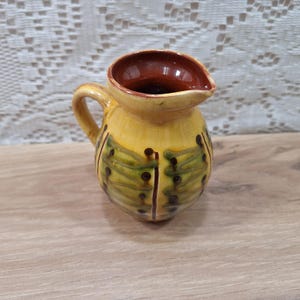 May include: A small, hand-painted ceramic pitcher with a yellow base and a brown interior. The pitcher features green and brown leaf-like designs and a curved handle. The spout is shaped for easy pouring. A decorative piece for the kitchen.