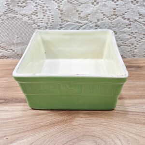 May include: A square, ceramic baking dish with a light green exterior and a cream-colored interior. The dish has a ribbed design around the sides and a white rim. The dish is empty and sits on a wooden surface.