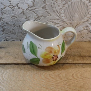 May include: A white ceramic pitcher with a rounded body and a handle. The pitcher is decorated with a yellow and orange floral design and green leaves. The pitcher is sitting on a wooden surface.