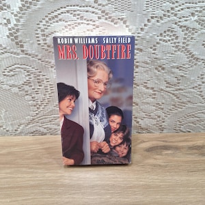 May include: A VHS tape for the movie "Mrs. Doubtfire" featuring Robin Williams and Sally Field. The cover art shows a woman in glasses and a floral sweater, with other characters peeking from behind a door. The title is in red.