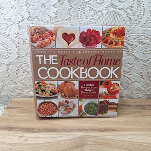 May include: A cookbook titled "The Taste of Home Cookbook" with various food images on the cover. The cover features images of prepared dishes, including a heart-shaped pastry, raspberries, and a roasted chicken. The book is on a wooden surface.