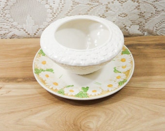 May include: White ceramic bowl with a woven pattern and a matching saucer with a daisy design. The saucer has a yellow rim and green leaves around the daisies.