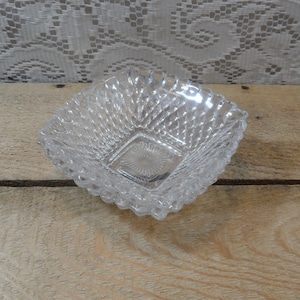 May include: A clear, square glass dish with a diamond pattern and scalloped edges. The dish has a square base with a starburst design. The glass is transparent, allowing light to pass through. The dish is likely vintage or antique.