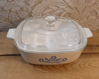 May include: A vintage white casserole dish with a clear glass lid and a clear glass knob. The dish has a square shape with rounded corners and blue floral designs. The dish has two white handles.