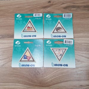 May include: Four Girl Scout Try-Its iron-on patches on teal backing. Each patch is triangular with a different design, including a ship, a baked good, a triangle with numbers, and various symbols. The patches are ready to be ironed on.