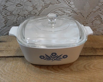 May include: A white square casserole dish with a clear glass lid. The dish has a blue floral design on the front and white handles. The lid has a clear knob. The dish is on a wooden surface.