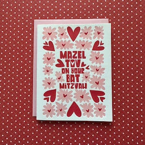 May include: A greeting card with the text "Mazel Tov on your Bat Mitzvah" in red. The card features pink flowers and red hearts on a white background, with a pink envelope. The background is red with white polka dots.