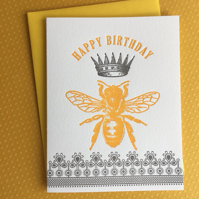 May include: A white card with the text "Happy Birthday" in black lettering. A black and white crown sits above a yellow bee with black markings. The card has a black and white decorative border.