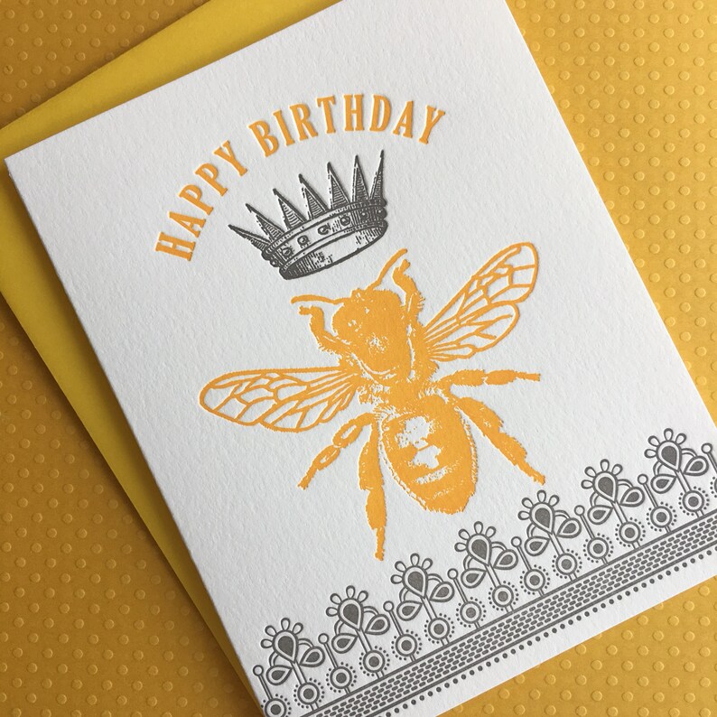 May include: A white card with the text "Happy Birthday" in black lettering. A yellow bee with a black crown on its head is in the center of the card. A black and white decorative border is at the bottom of the card.