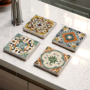 Può includere: Quattro sottobicchieri quadrati in ceramica con intricati disegni floreali colorati. Ogni sottobicchiere presenta un motivo unico con sfumature di blu, giallo, arancione e verde. I sottobicchieri sono disposti su un piano di lavoro bianco, mostrando il loro fascino decorativo.